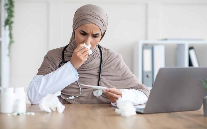Sick young muslim woman in hijab doctor sneezing nose and looking at thermometer, checking fever, sitting at workdesk with laptop in office at clinic, got sick during COVID-19 pandemic, copy space